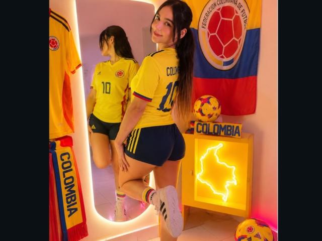 MindyLove in a yellow soccer jersey with "COLOMBIA" and number 11, showing her ass in tight shorts, standing with one leg up, reflected in a mirror.