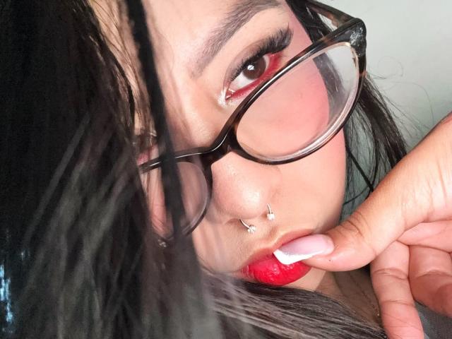 ChloeMoretti with red lipstick, black glasses, and nose piercings, biting her finger, showing off glossy pink nail, and dark braided hair, looking up with red eyes.