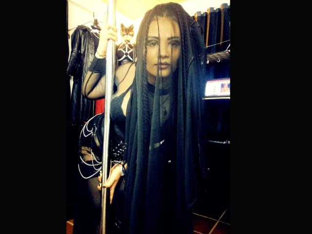 Die Göttin Mafdet mit langen, schwarzen Dreadlocks, die einen Stab hält, trägt ein schwarzes Lederkleid mit Spikes und blickt direkt in die Kamera, in einem schwach beleuchteten Raum..