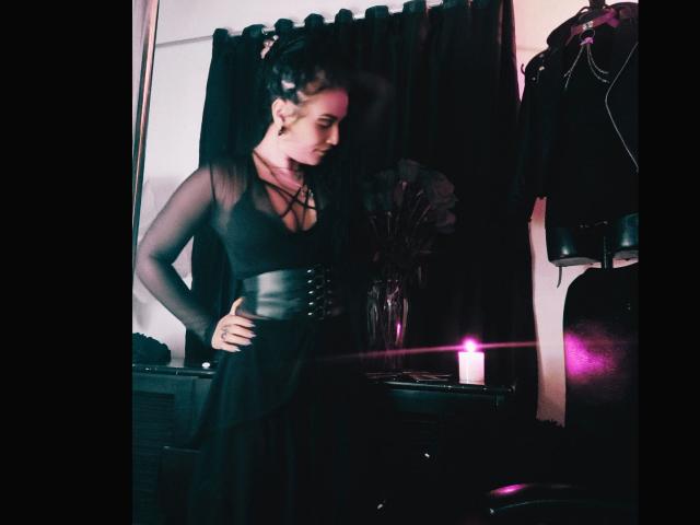 La déesse Mafdet, vêtue d'une robe noire transparente aux manches fines et à la taille ajustée comme une corset, se tient debout, une main sur la hanche, le regard baissé, dans une pièce faiblement éclairée avec une bougie..
