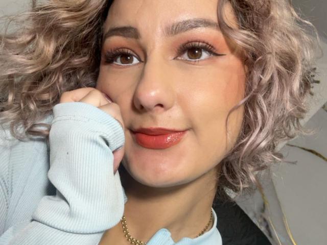 Flora Saenz, met krullend, roze-blonde haar, bruine ogen, oranje eyeliner, glanzende rode lippen, draagt een lichtblauwe trui en raakt haar wang aan met een gouden ketting..