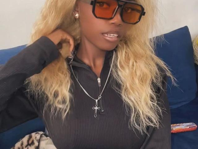 EbonyAngel: pelle marrone scura, capelli biondi ondulati, occhiali da sole neri, felpa nera a coste, collana con croce d'argento e piccoli orecchini, con un leggero sorriso..