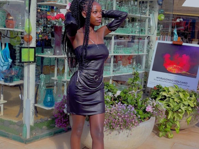 EbonyAngel, atractiva con un vestido negro ajustado y brillante, de manga larga y con un cinturón de cadena, está de pie con las manos en las trenzas, mostrando sus curvas y su piel oscura y suave..