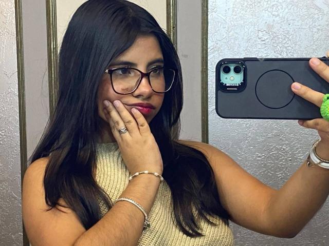 Celeste Morel, com cabelo escuro, óculos, piercing no nariz e batom rosa, segurando um telefone, vestindo uma blusa bege sem mangas, com a mão no rosto, um anel no dedo e uma pulseira no pulso..