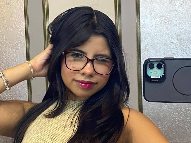 Celeste Morel tem cabelo escuro, óculos, batom rosa e uma blusa bege. Ela está piscando com um olho, com a mão no cabelo, e tem uma pulseira no pulso. Um celular está no fundo..