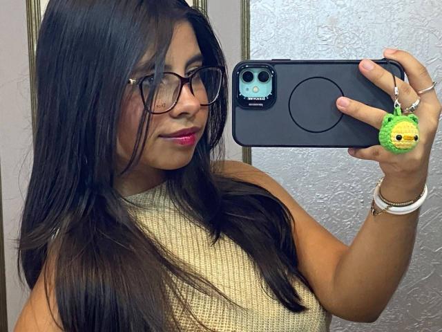 Celeste Morel, com cabelos longos e escuros, óculos, batom rosa, blusa bege sem mangas, segurando um telefone preto com uma capa de pássaro verde, pulseira branca, sorrindo, selfie no espelho do banheiro..