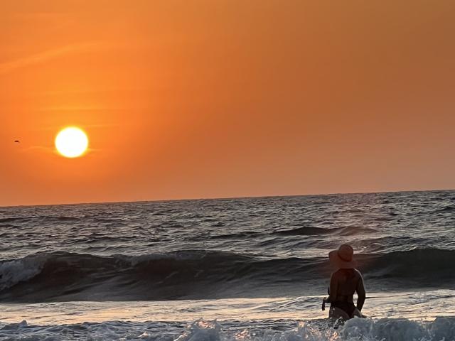 SashaPolet stands in the ocean, wearing a wetsuit and a straw hat, facing a sunset with a glowing orange sky and a bright sun near the horizon.