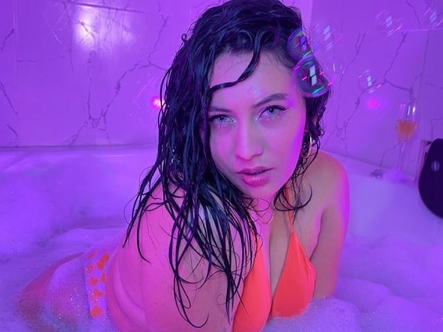 Amy Ross, avec ses cheveux noirs mouillés, portant un bikini orange, dans une baignoire éclairée en violet, regardant l'appareil photo avec des yeux pétillants, ses petites seins et ses fesses partiellement couverts de bulles..