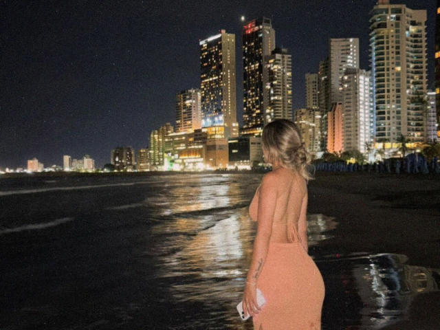 SashaPolet stands on a dark beach at night, wearing an orange, backless dress that shows her ass and holding a white phone, with city skyscrapers in the background.