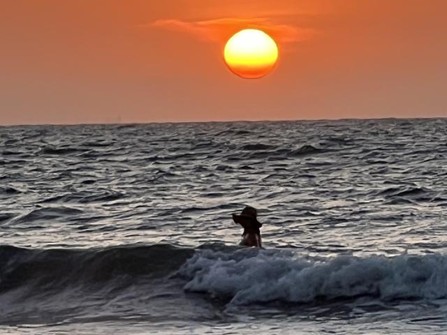 SashaPolet está no oceano, surfando uma pequena onda, com os braços levantados, o sol se pondo no céu laranja, água ao redor da cintura, e ondas em primeiro plano..