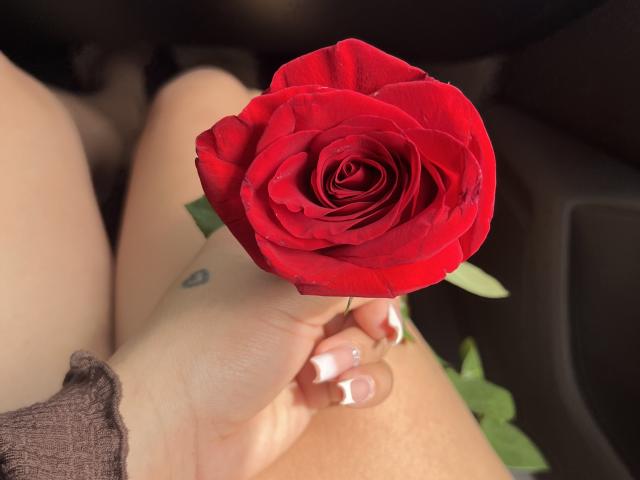 A mão de SashaPolet segurando uma rosa vermelha, exibindo unhas bem cuidadas com pontas brancas, sua coxa e uma pequena tatuagem visíveis, em um ambiente escuro, semelhante a um carro..