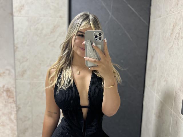 SashaPolet, com cabelos loiros e ondulados, vestindo um vestido preto com um decote profundo em V, exibindo seus seios grandes, segurando um telefone prateado, sorrindo, em pé em um quarto com piso de azulejos..