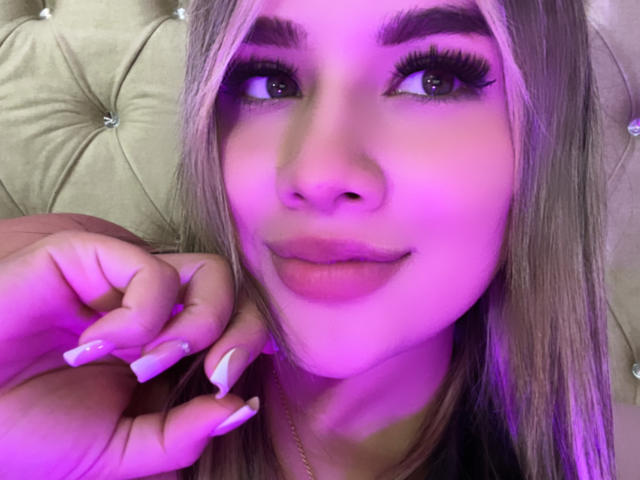 SashaPolet's Gesicht mit violetter Beleuchtung, langes blondes Haar, dicke Augenbrauen, grüne Augen, lange Wimpern, nacktrosa Lippen, gepflegte Nägel, Hand in der Nähe des Gesichts, zur Seite blickend, lächelnd..