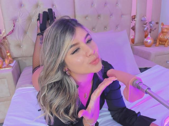 SashaPolet with blonde hair, black fishnet gloves, and a black shirt, pouts her pink-lipsticked lips, touching a microphone with her right hand, legs up, bed in background.