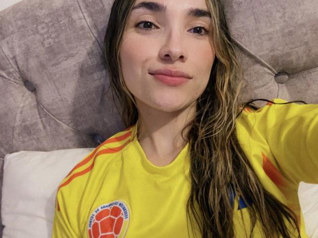 AdharaCooperr with wet brown hair, yellow soccer shirt, and red stripes, smiling at camera, lying on gray tufted headboard, showing cute face and pink lips.