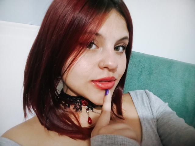 Sophie Hill mit roten und braunen Haaren, Nasenring und schwarzem Choker mit rotem Stein, die ihre glänzlichen, rosafarbenen Lippen mit einem violetten Nagellack berührt, trägt ein graues Oberteil..