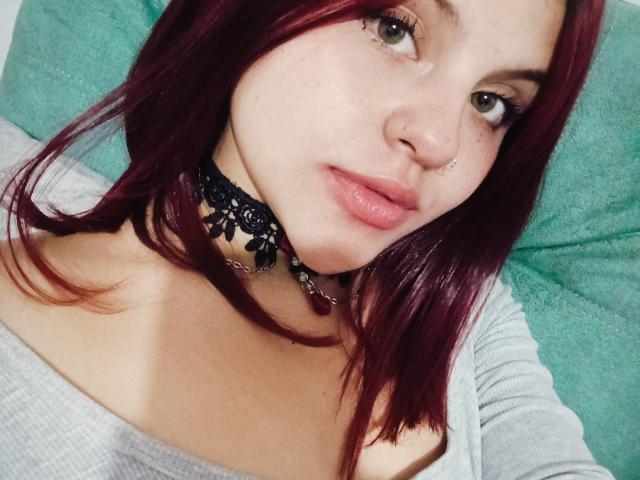 Sophie Hill, avec ses cheveux roux, son anneau de nez et son collier en dentelle noire, porte un haut gris et regarde vers le haut avec ses yeux verts et ses lèvres roses. Ses seins sont légèrement visibles..