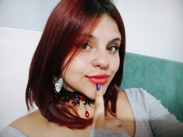 Sophie Hill, con i capelli rosso-marrone, un anello al naso, rossetto viola, una maglia grigia, una collana nera in pizzo con una pietra rossa e unghie viola, tocca delicatamente le labbra con un dito..