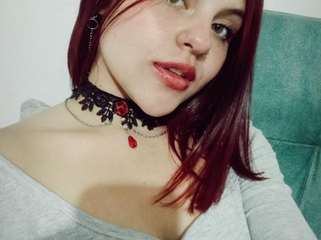 Sophie Hill, con cabello rojo, aro en la nariz, collar de encaje negro con una rosa roja, labios rojos y brillantes, vestida con una blusa gris claro, sentada en una silla verde, mirando ligeramente hacia la derecha..