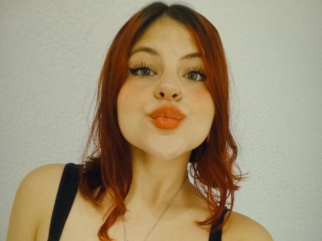 Sophie Hill tem cabelo ruivo, um piercing no nariz e batom laranja. Ela está vestindo uma blusa preta com um colar de prata e está fazendo beicinho..