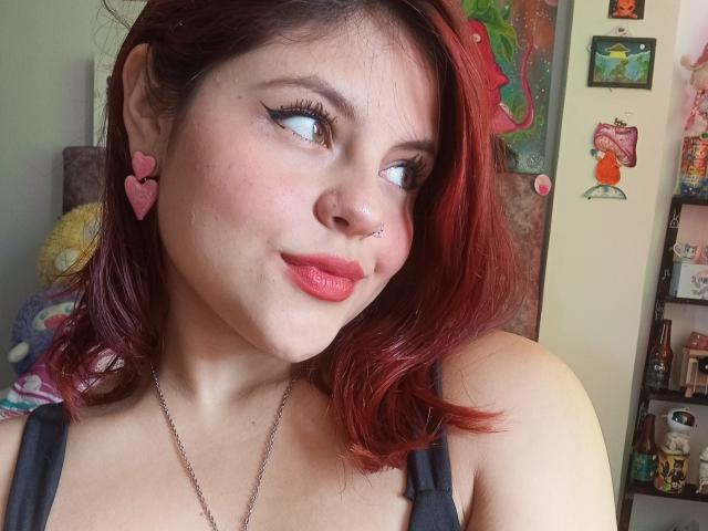 Sophie Hill hat rotes Haar, herzförmige Ohrringe in Rosa, einen Nasenring und roten Lippenstift, sie schaut zur Seite mit einem leichten Lächeln und trägt ein schwarzes Tanktop, das ihre Brüste zeigt..