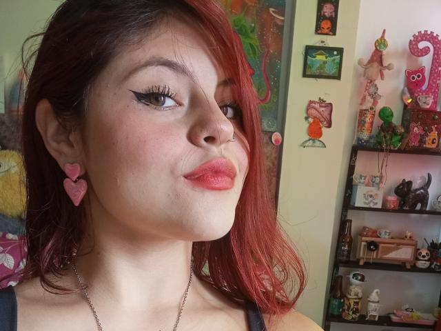 Sophie Hill, met rood haar, zwarte eyeliner, roze hartvormige oorbellen, een neusring en roze lippenstift, glimlacht lichtjes en laat haar borsten zien in een selfie, terwijl ze een zwarte top draagt..