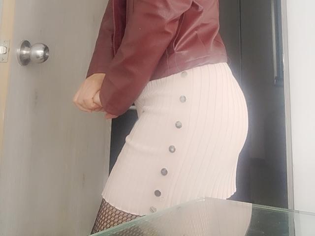 CamilleRiver indossa una giacca di pelle color marrone scuro, una gonna bianca con bottoni neri, calze a rete e si trova in posizione tale che il sedere e una mano sono visibili..