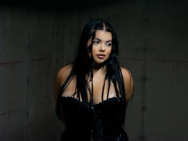 LaraJames heeft lang zwart haar, blauwe ogen en draagt een zwarte corset die haar grote borsten laat zien. Ze ziet er serieus uit met een licht geopende mond..