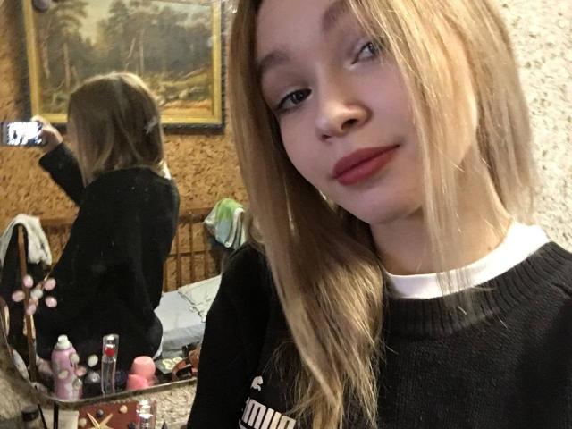 AnnamaeSvehla with long blonde hair, wearing a black sweater, holding a phone, mirror reflecting her back, red lipstick, white collar, cluttered makeup table in the foreground.