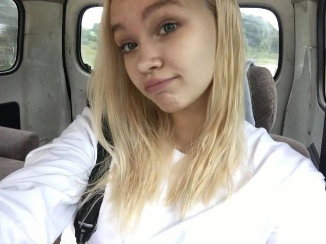 AnnamaeSvehla with blonde hair, blue eyes, and a slight smile, wearing a white shirt, sitting in a car, her small tits barely visible under the seatbelt.