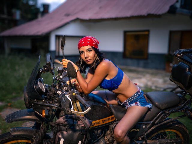 EvelinLuna with red bandana, blue bra, and star-spangled shorts, leaning on a Yamaha Enticer motorcycle, her tits barely covered, looking seductive, outdoor evening, motorcycle in front.