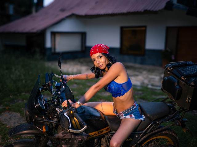EvelinLuna mit rotem Bandana, blauem Spitzen-BH und kurzen Jeansshorts, fährt eine schwarze Motorrad und zeigt ihr trainiertes Gesäß sowie einen gelben Oberschenkelgurt, in einer grünen, im Dämmerlicht gelegenen Umgebung..