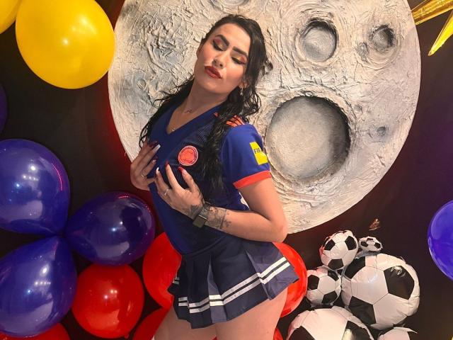 DemiMistral with long black hair, wearing a blue soccer jersey, red and yellow patches, and a short skirt, touching her big, round tits, surrounded by colorful balloons and a soccer ball.