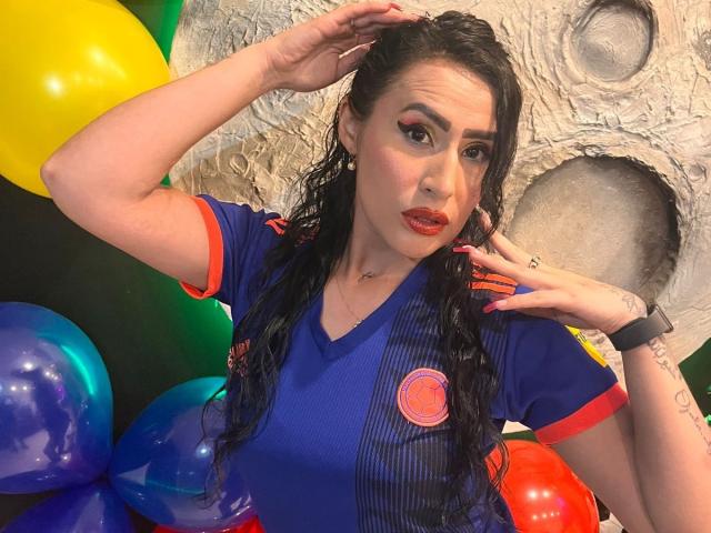 DemiMistral, avec ses cheveux noirs et humides, portant un maillot de foot bleu avec des détails rouges, des lèvres rouges et une expression sérieuse, touchant sa tête avec des ballons colorés en arrière-plan..