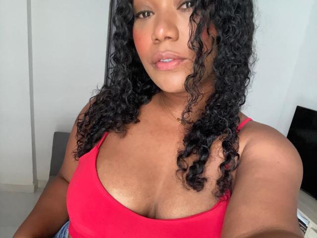 AvaJones69 with curly black hair, red tank top, and visible large boobs, has a serious expression, light brown skin, and pink lipstick, sitting in a bright room.