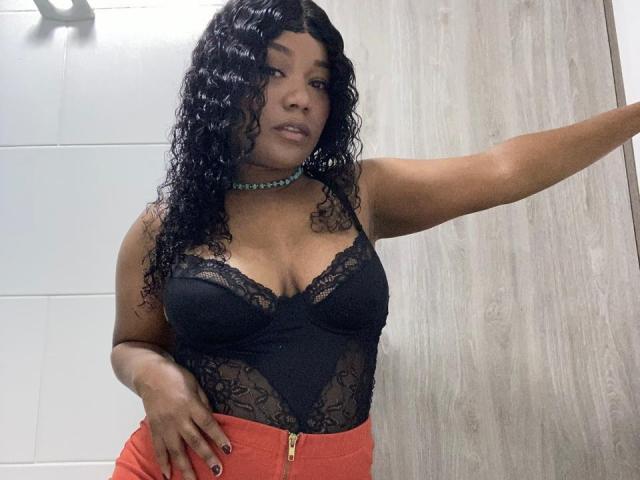 AvaJones69 with curly black hair, wearing a black lace bodysuit showing her boobs, red pants, and a green bead necklace, standing against a wooden door.