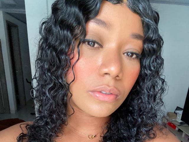 AvaJones69 with curly black hair, brown skin, and pink lips, wearing a gold heart necklace, staring into the camera with slightly parted pink lips.