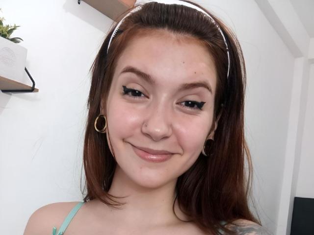 AliciaBach hat helle Haut, braune Augen und ein leichtes Lächeln. Sie trägt einen weißen Haarreif, Ohrringe im Stecker-Stil und hat eine Nasenring. Ihr Make-up umfasst einen dicken schwarzen Eyeliner..
