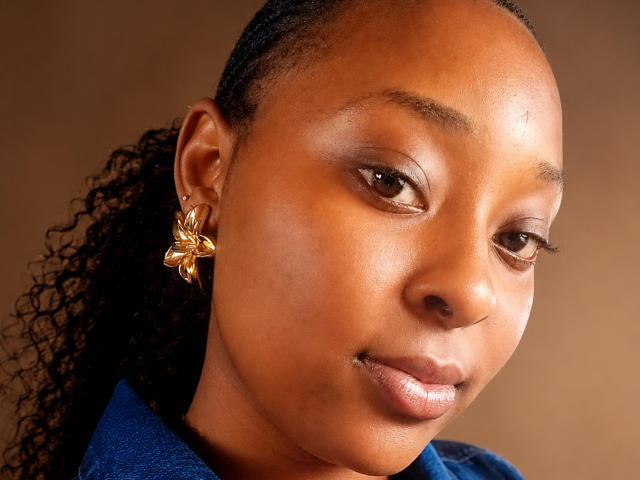 Le visage de TiffanySwit, avec sa peau brune, ses cheveux noirs bouclés, ses boucles d'oreilles dorées en forme de fleurs et son maquillage discret. Elle porte un haut bleu et regarde légèrement vers la droite..