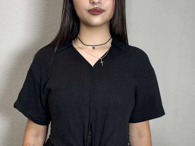 MaeCopass met lang zwart haar, gekleed in een zwarte top, met een kruisbeeldketting en een zwarte choker, die haar borsten laat zien, met rode lippenstift, staand voor een effen grijze muur..