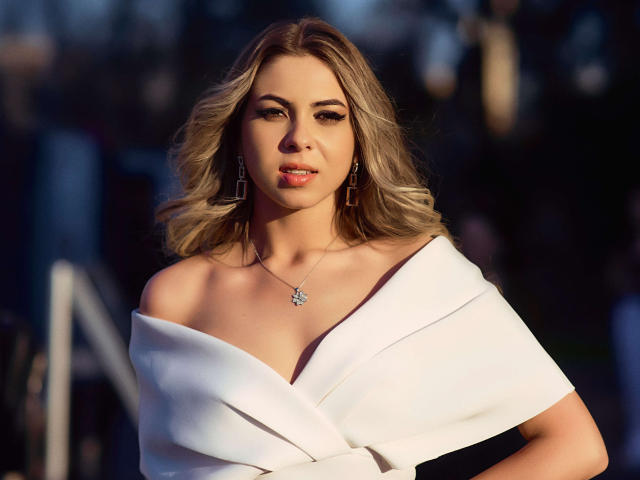 AylinRoze mit welligem, blondem Haar, einem weißen Kleid mit Schulterpartie, kleinen Brüsten und einer zarten Halskette, die selbstbewusst und verführerisch in einem sonnendurchfluteten, unscharfen Hintergrund wirkt..