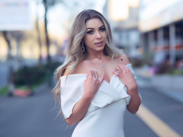 AylinRoze met lang blond haar, een witte jurk met open schouders, handen op de borst, een kleine harttatoeage in de buurt van het sleutelbeen, kruis oorbellen, een subtiele glimlach, een onscherpe straat op de achtergrond..