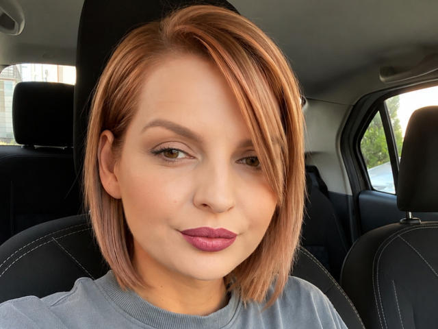 DouceAmelia con un corte de pelo corto, castaño rojizo, piel clara, maquillaje sutil, lápiz labial morado, camisa gris, asiento de coche negro, ligera sonrisa, mirando a la cámara, luz diurna, expresión suave..