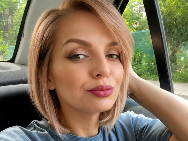 DouceAmelia heeft een schattig gezicht met roze lippenstift, groene ogen en een blond kort kapsel, ze zit in een auto, draagt een blauw shirt en heeft een tatoeage op haar rechterarm..