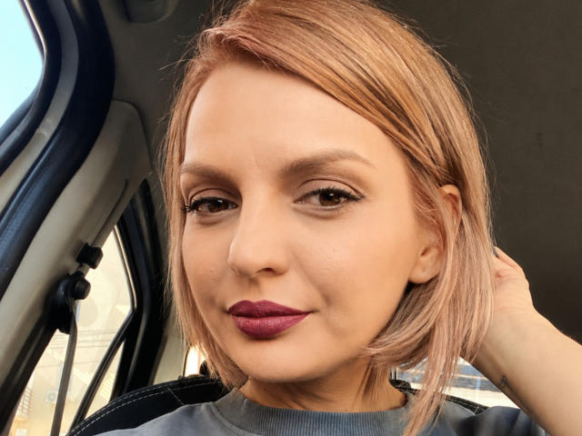 DouceAmelia, con piel de tono marrón medio, cabello lacio de color rosa-marrón, delineador de ojos oscuro y lápiz labial morado, sentada en un coche, con la mano en el pelo, sonriendo suavemente. Los senos no son visibles..