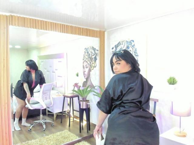 ViviWollds in a black satin robe, facing mirror, showing her ass, reflection shows her standing with a white chair, wooden floor, and a green plant.