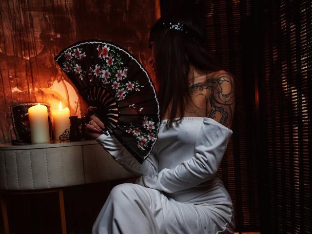 KayaBelle, avec des tatouages sur le dos, tenant un éventail fleuri, vêtue d'une robe blanche à une épaule, est assise près de bougies allumées, son postérieur étant à peine visible. Pièce sombre, avec des ombres..