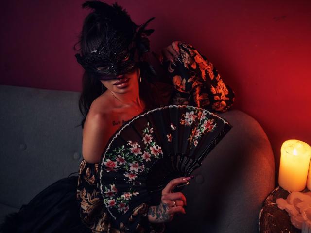 KayaBelle, com cabelo escuro, máscara de renda preta, leque preto com flores e braço tatuado, sentada em um sofá cinza, com luz vermelha, vestindo um vestido preto e com uma pena preta no cabelo..