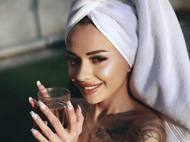 KayaBelle with a white towel on her head, holding a glass of brown liquid, smiling with glossy skin, long white nails, and visible tattoos on her chest.