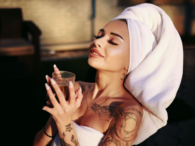 KayaBelle with a white towel wrapped around her head, holding a glass of brown liquid, showing off tattoos on her arms and chest, smiling with eyes closed.